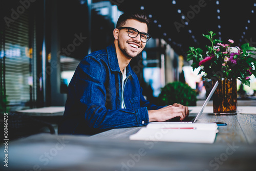 Portrait of cheerful male copywriter in eyewear typing text on laptop computer for publication satisfied with remote job, smiling hipster guy blogging in social networks chatting with followers online
