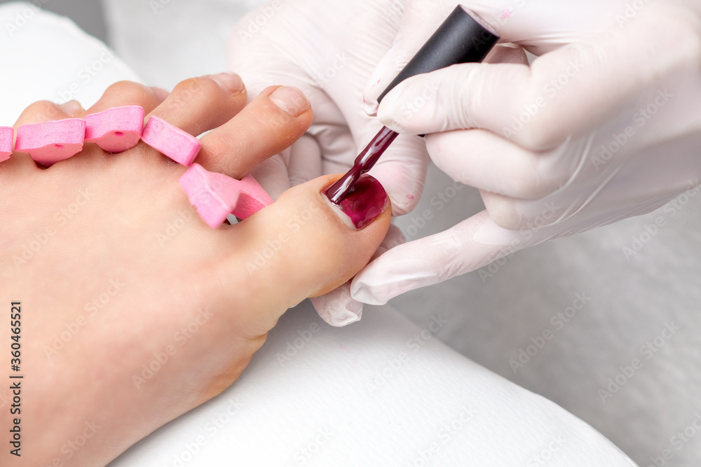 Hands in protective gloves painting red nail polish on female toenail in beauty salon Hands in protective gloves painting red nail polish on female toenail in beauty salon