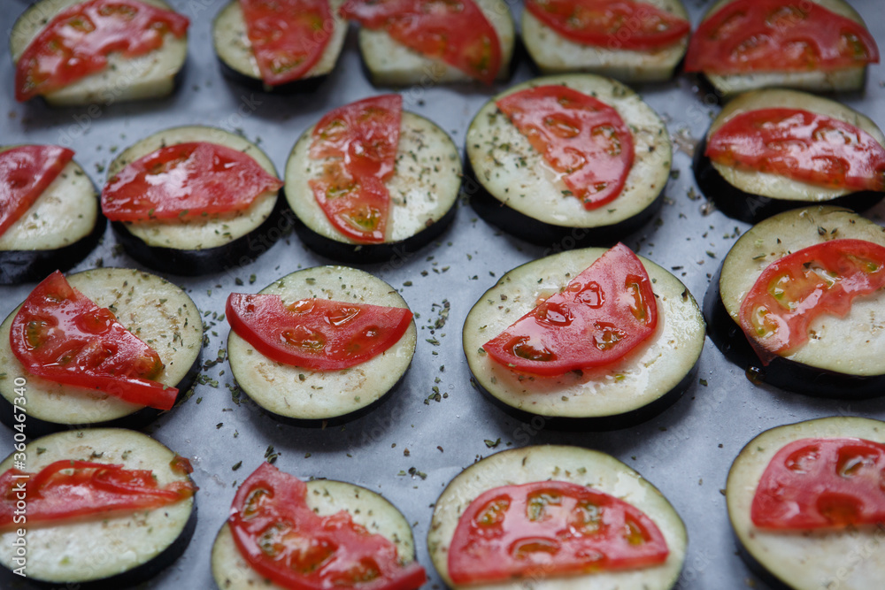 isolated filled frame close up shot of uncooked raw sliced aubergine eggplant with thin tomato slices on top sprinkled with dry green basil and oregano herbs, lying on a white baking paper background isolated filled frame close up shot of uncooked raw sliced aubergine eggplant with thin tomato slices on top sprinkled with dry green basil and oregano herbs, lying on a white baking paper background