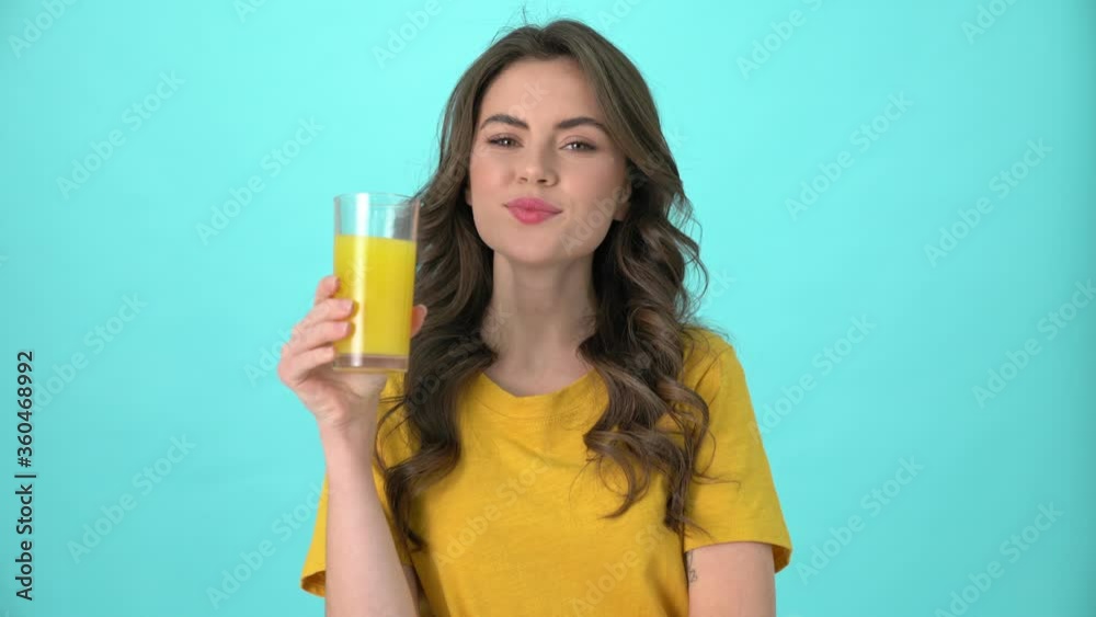 A friendly young woman wearing a yellow t-shirt is drinking fresh juice and showing thumb-up sign standing isolated over blue background A friendly young woman wearing a yellow t-shirt is drinking fresh juice and showing thumb-up sign standing isolated over blue background