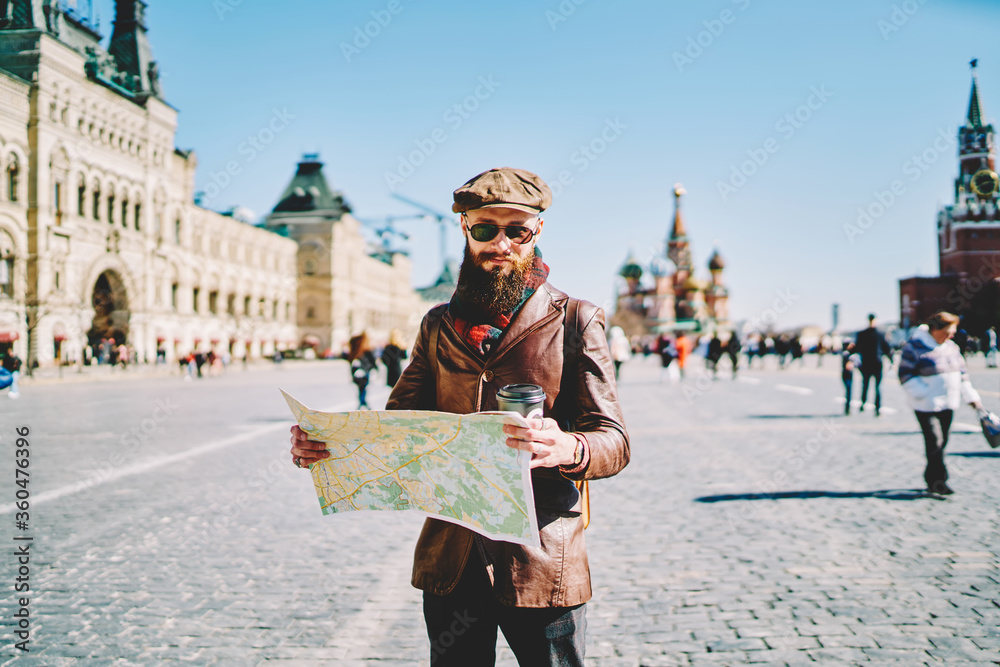 Obraz premium Young caucasian traveller dressed in trendy clothing searching location on paper map, stylish hipster guy trying find new sightseeing for discover while walking on Moscow Red Square during vacation