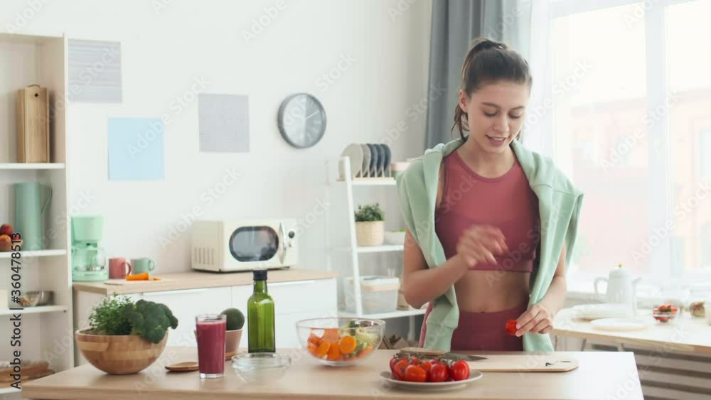Medium shot of young Caucasian sportswoman standing at kitchen table, making vegetable salad while recording it on camera for her blog Medium shot of young Caucasian sportswoman standing at kitchen table, making vegetable salad while recording it on camera for her blog