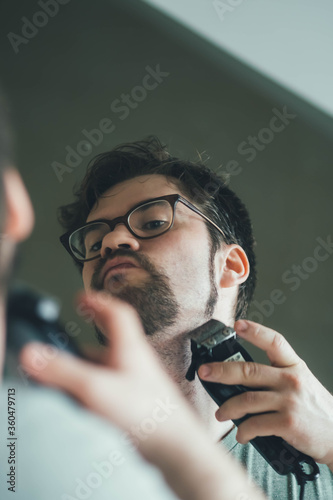 a handsome young man with a thick beard and glasses is illuminated on one side by a soft light. fashionable beard, happy. reflection in the mirror. irritation after shaving. shaves three times