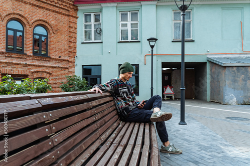 a hipster is sitting on a bench with a phone in his hands in a new block, a white man in a hat sat down to rest, a stylish guy