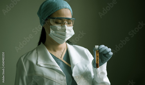 Medical lab worker looks at blood test tube with Covid 19 inscription. Young Asian woman wearing uniform, gloves and goggles on gray background. Healthcare concept. Toned image. photo