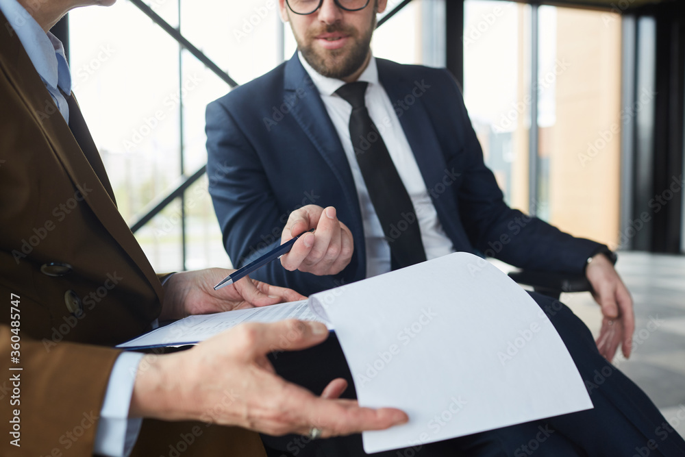Close-up of businessman pointing at business contract and discussing details with businesswoman while they sitting at office Close-up of businessman pointing at business contract and discussing details with businesswoman while they sitting at office