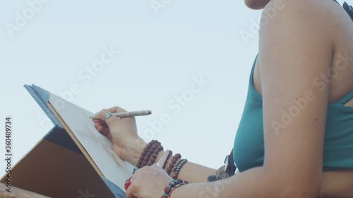 Low angle view of girl drawing sketch on paper clipboard outdoors on blue sky background. Faceless artist working on picture on summer warm day macro detail. Art workshop creative process photo