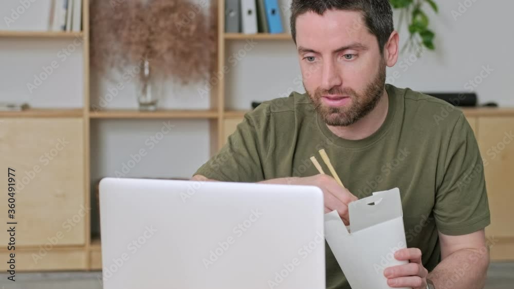 A smiling positive young man is watching something funny on his laptop while sitting at the table in the office during dinner A smiling positive young man is watching something funny on his laptop while sitting at the table in the office during dinner