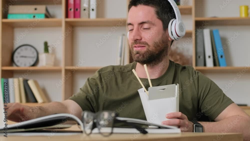 A positive pleased young man is watching something interesting on his laptop during dinner sitting at the table in the office A positive pleased young man is watching something interesting on his laptop during dinner sitting at the table in the office