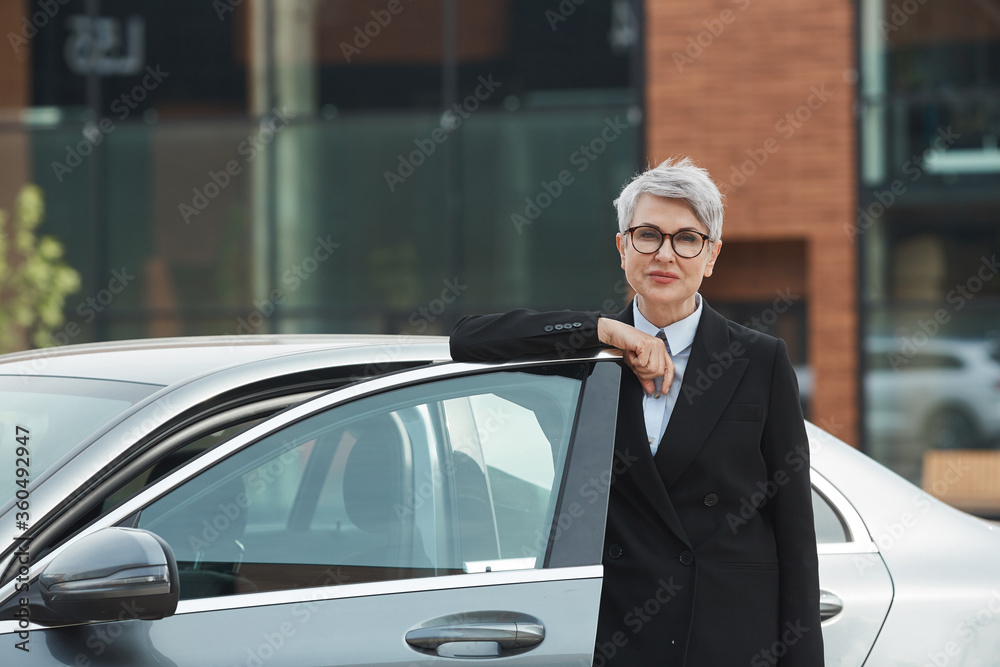 Portrait of mature businesswoman in eyeglasses standing near the car and looking at camera outdoors
