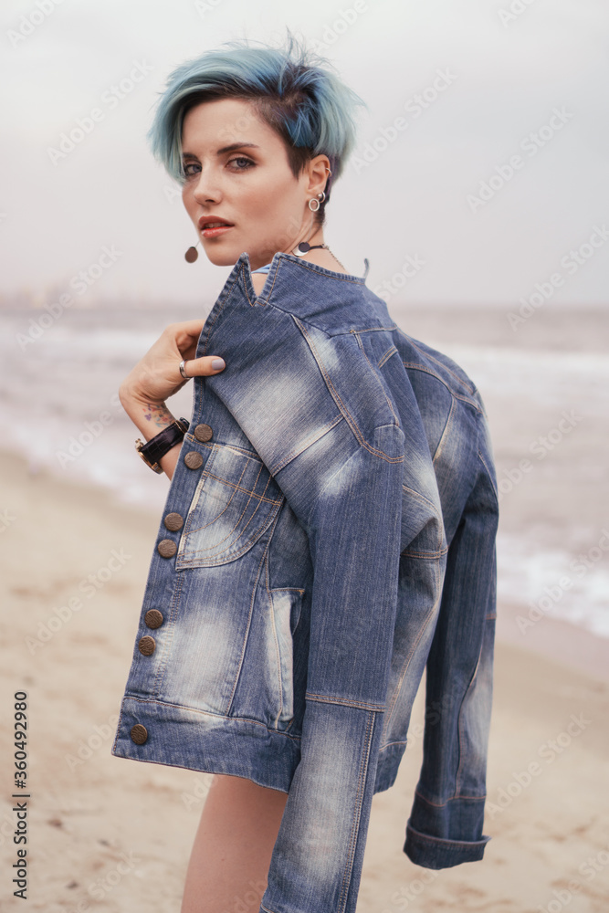 Beautiful young girl in a blue swimsuit and a denim jacket on her shoulders posing on the coast