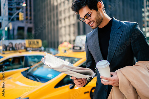 Smiling successful entrepreneur in elegant suit satisfied with article in daily press standing on avenue with blank coffee cup, happy businessman reading newspaper waiting for yellow cab on Manhattan.