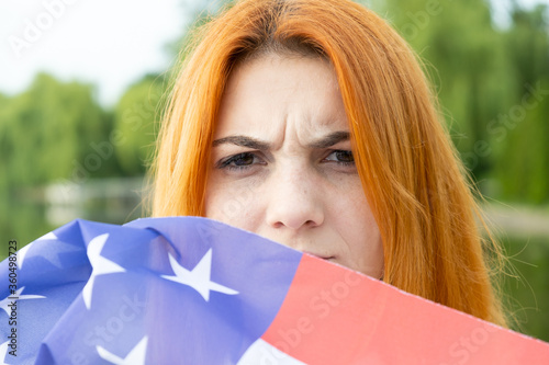 Portrait of angry red haired girl hiding her face behind USA national flag.