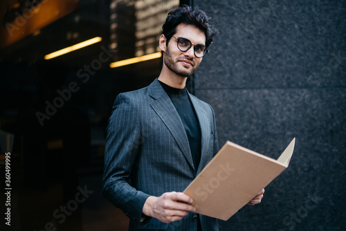 Half length portrait of successful young executive manager dressed in elegant wear smiling at camera while holding file with financial documents standing outdoors.Positive banker with folder in hands