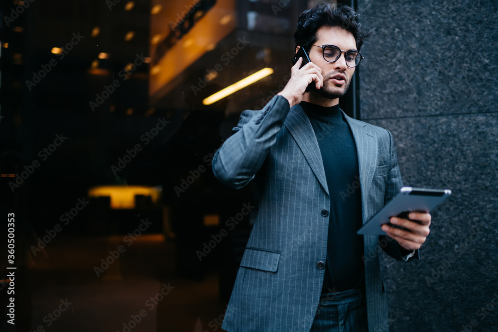 Serious male entrepreneur in elegant outfit having telephone conversation confirming booking via tablet, serious confident businessman making mobile phone call reading information from touchpad.