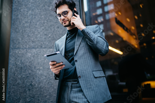 Serious male entrepreneur in elegant suit talking on mobile phone reading information from digital tablet, confident businessman making booking calling to service operator holding portable pc.