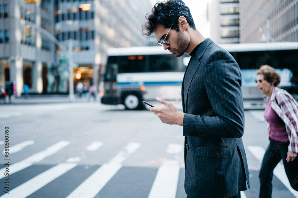 Side view of pensive young entrepreneur in stylish suit reading financial news in mobile application crossing zebra in downtown of metropolis.Confident proud CEO checking banking account on cellular Side view of pensive young entrepreneur in stylish suit reading financial news in mobile application crossing zebra in downtown of metropolis.Confident proud CEO checking banking account on cellular