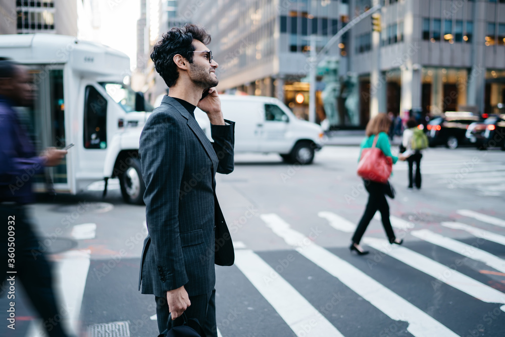 Side view of sucessful young stylish owner making telephone conversation strolling across the street of downtown, smiling man in trendy suit and eyewear talking on cellphone using roaming connection.