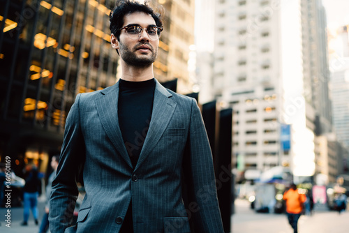 Portrait of handsome owner of business dressed in stylish trendy suit and spectacles looking at camera outdoors, young serious caucasian man in fashion outfit walking on avenue in centre of the city. © BullRun