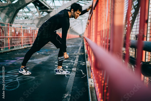 European sportsman in trendy tracksuit stretching on city bridge before start running across street urbanity, young jogger doing workout outdoors during listening radio via electronic headphones © BullRun