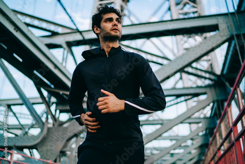 Muscular jogger in sport clothing running on city bridge during morning time, active european male athlete jogging outdoors and looking away during hard cardio training for making body strength