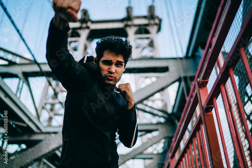 Concentrated young professional boxer dressed in black tracksuit is engaged in sport in urban setting to achieve goal.Concentrated fit man in active wear boxing outdoors during workout