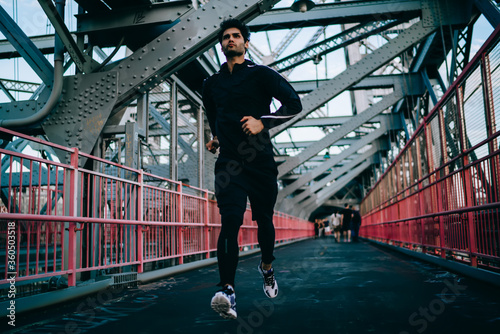 Motivated male jogger doing cardio to maintain sporting form in urban setting.Young athlete dressed in black tracksuit running on bridge.Fit man in active wear and sneakers training outdoors © BullRun