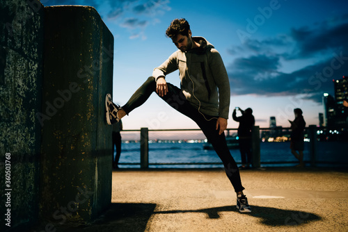 Sportsman dressed in tracksuit doing stretching while enjoying music during evening workout in urban setting.Young man in sport wear doing exercise training outdoors while listening audio in earphones