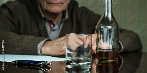 aged man looks at whiskey bottle and glass sitting at wooden table with sheet of paper