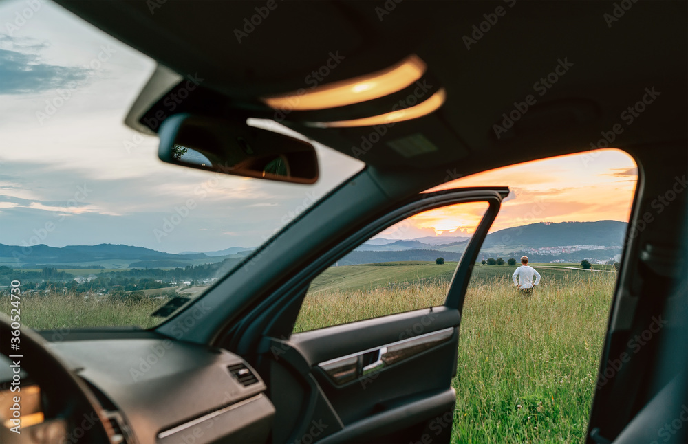 Young man standing in high grass and enjoying the sunset sky colors through the open car dor view. Traveling by car concept image. Young man standing in high grass and enjoying the sunset sky colors through the open car dor view. Traveling by car concept image.