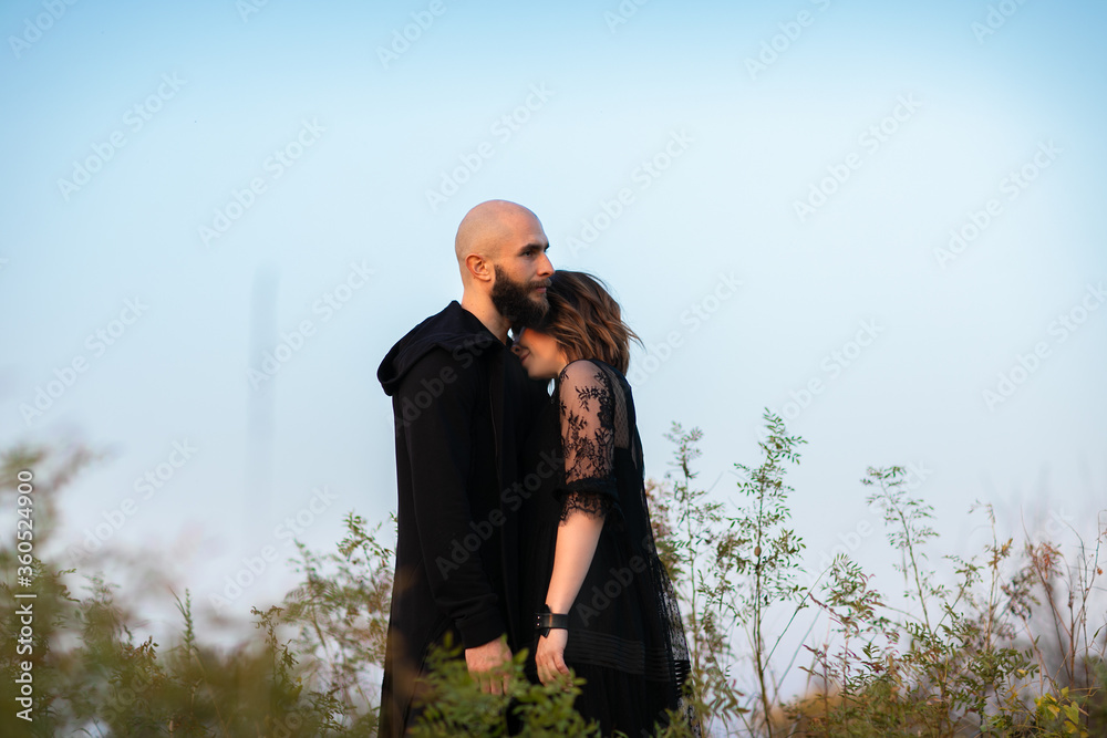 Portrait brunette woman and bald headed man with beard in black clothes posing on a hill. Sky background.