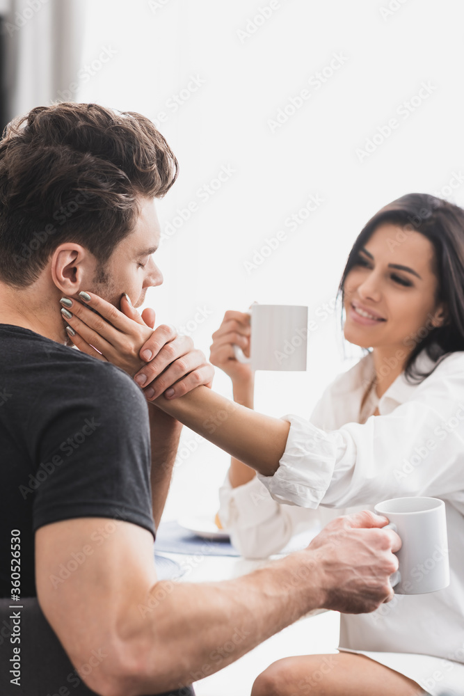 Selective focus of smiling woman touching face of boyfriend while drinking coffee in kitchen Selective focus of smiling woman touching face of boyfriend while drinking coffee in kitchen