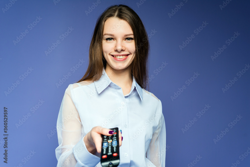 A happy woman in a blue shirt watches movies and TV shows on television. Beautiful girl in the studio on a lilac background switches channels using a remote control A happy woman in a blue shirt watches movies and TV shows on television. Beautiful girl in the studio on a lilac background switches channels using a remote control
