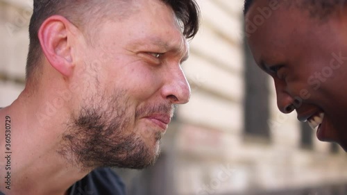 Outdor close portrait of young students. Side view. Black man and white man hugging and smiling. Equality, friendship complicity concept. No racism
