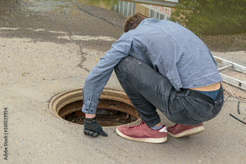 Man carefully looks at  open Manhole. Squatting bent over. View from the back. Concept of eliminating accident, laying communications, communication wires. © Sergei Tim