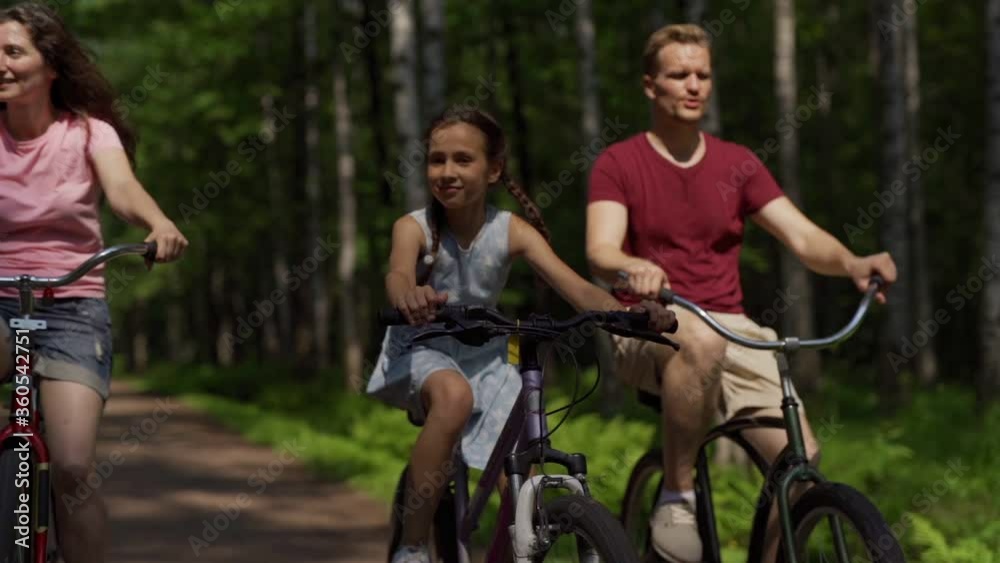 Tilt up slow motion shot of happy family of father, mother and little daughter enjoying riding bikes down trail in forest and talking on the move Tilt up slow motion shot of happy family of father, mother and little daughter enjoying riding bikes down trail in forest and talking on the move