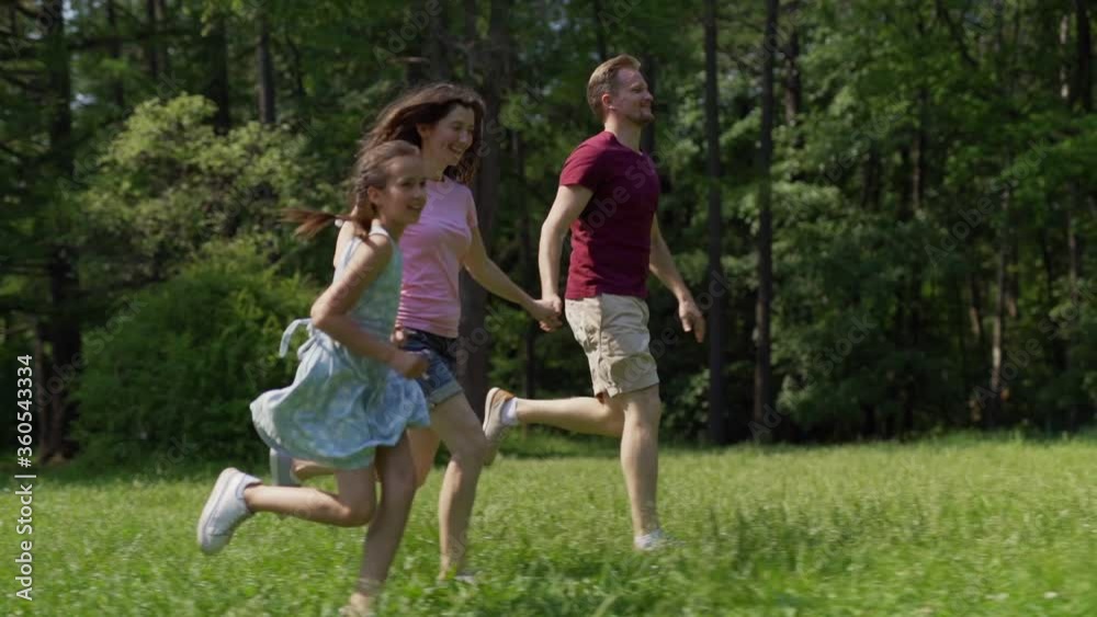 Side view slow motion shot of happy family of father, mother and little daughter running on green grass holding hands in summer park Side view slow motion shot of happy family of father, mother and little daughter running on green grass holding hands in summer park