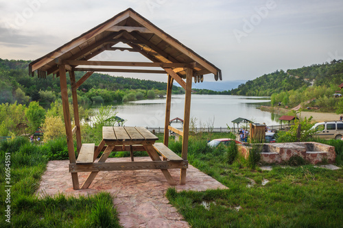 Wooden pergola in green grass near the lake in forest landscape. Picnic area in beautiful nature.
