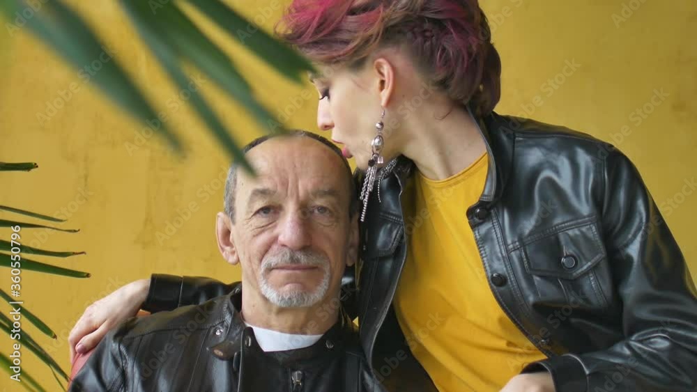 Young wife and senior husband are sitting on the armchair, woman in yellow dress and black leather jacket with dark pink hair is kissing her lover in a bald head. The couple with age difference