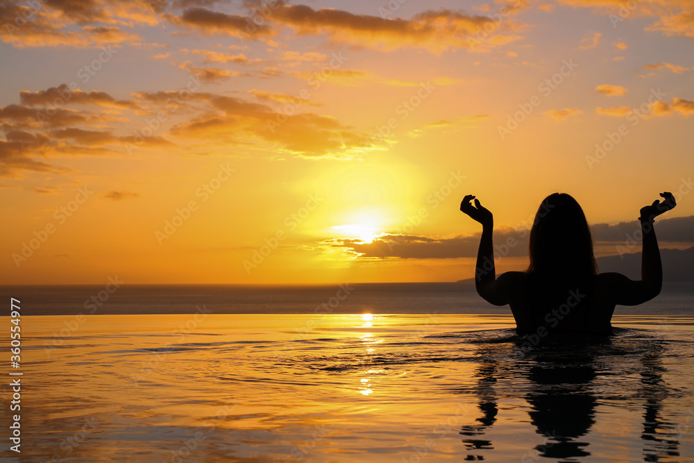 the girl is standing with her back to the pool, her hands folded as if for an Indian dance, against the background of the setting sun in the ocean. Gia de Isora. Tenerife. Spain