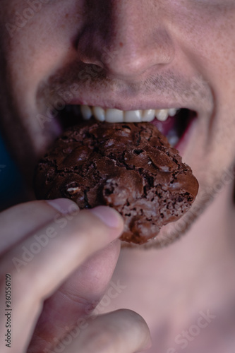 A detailed close-up shot of the mouth of a young handsome man who bites a cookie, holding it in his hand on a home background. Man eating chocolate cookie with peanut, white teeth, close up face.