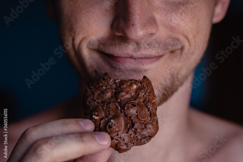 A detailed close-up shot of the mouth of a young handsome man who bites a cookie, holding it in his hand on a home background. Man eating chocolate cookie with peanut, white teeth, close up face.