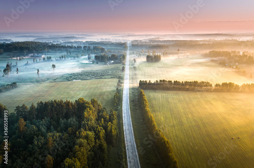 Highway going through meadow in mist surrounded by forest. Aerial view over picturesque landscape in fog at sunrise. 
