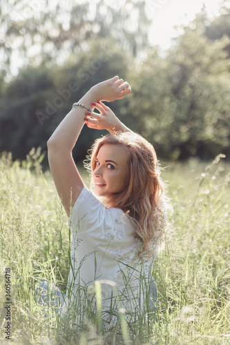 Beautiful blonde girl in a white T-shirt resting in a field with green tall grass, turns over her shoulder and stretches