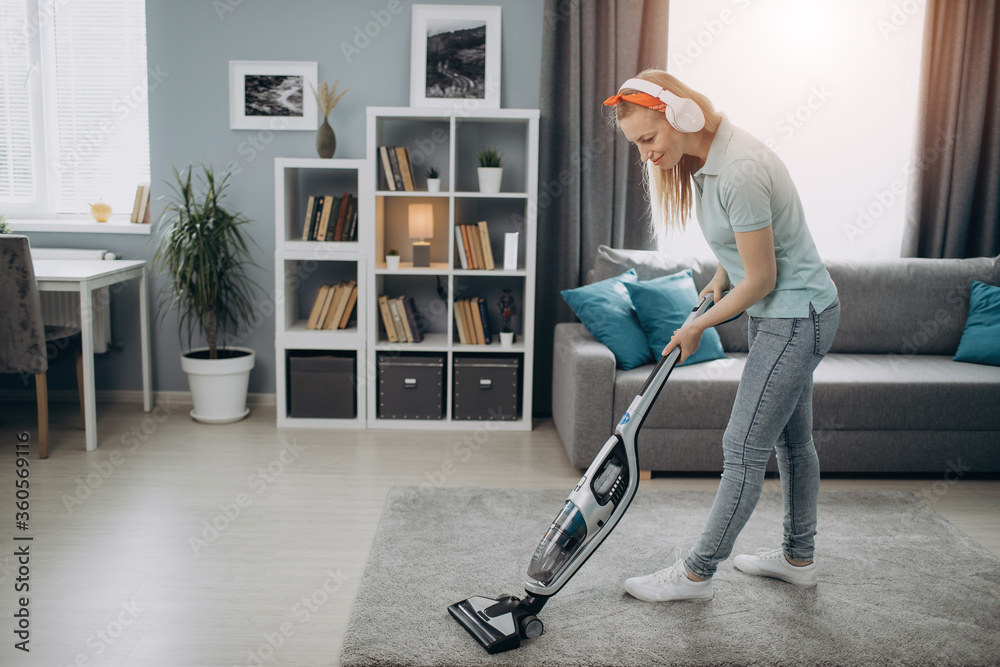 Full length portrait of beautiful woman in domestic clothing cleaning carpet with modern vacuum at home. Housewife with blond hair listening music in wireless headphones while doing household duties