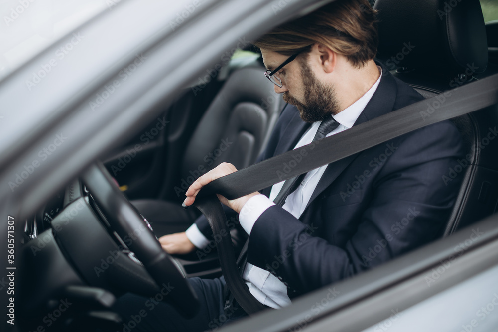 Mature bearded man dressed in formal suit sitting in car and putting on seatbelt before driving. Professional businessman protecting himself on road.