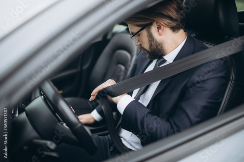 Mature bearded man dressed in formal suit sitting in car and putting on seatbelt before driving. Professional businessman protecting himself on road. © Kuz Production