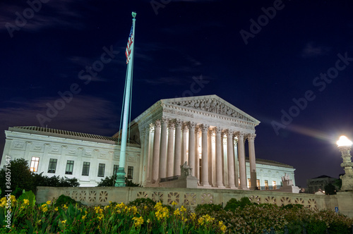 American flag flying on pole at night in front of the granite and marble greek style United States Supreme Court with yellow flowers in the foreground and clear sky and architecture lights