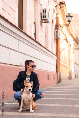 Beautiful caucasian girl with sunglasses squats on the street next to her dog breed American Staffordshire Terrier