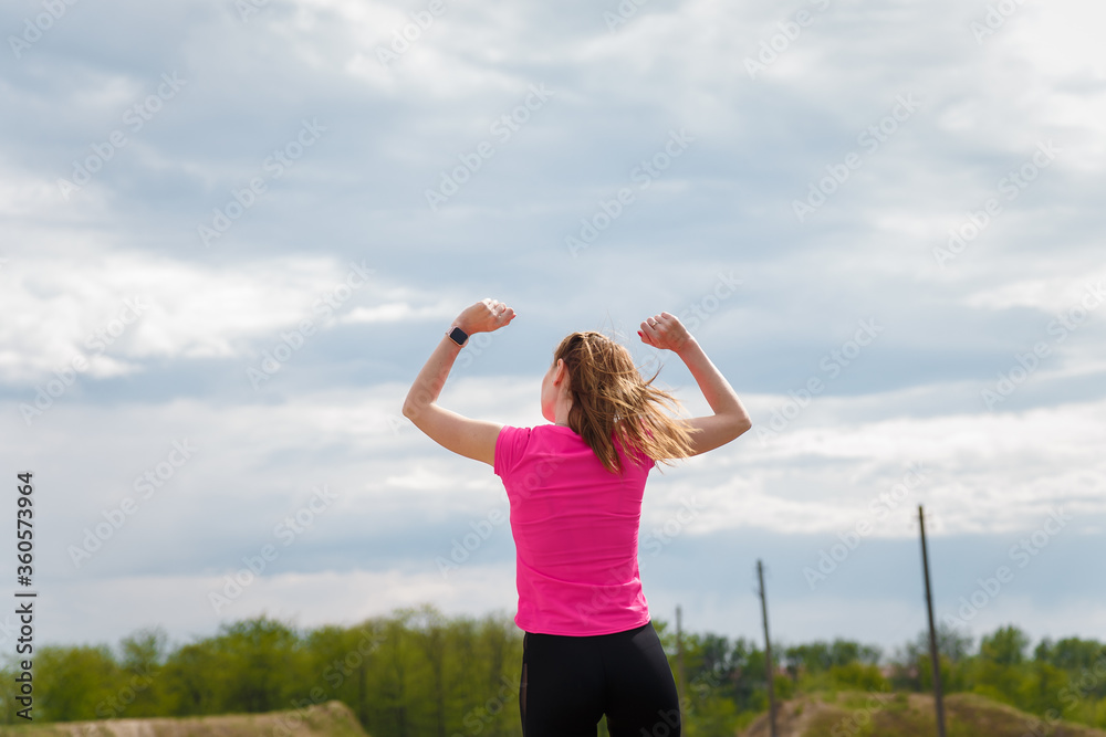 Young happy woman in sportswear standing on hill back to camera with arms up to sky. Fitness, healthy way of life, wellbeing, freedom, mental health, triumph, winning concept. Young happy woman in sportswear standing on hill back to camera with arms up to sky. Fitness, healthy way of life, wellbeing, freedom, mental health, triumph, winning concept.